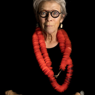 Kirsten Sonne photographed against a black background wearing a large red necklace made from many plastic disks.