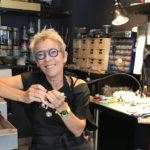 Kirsten Sonne sat in the middle of the frame, in her workshop. Wearing black clothes and apron.